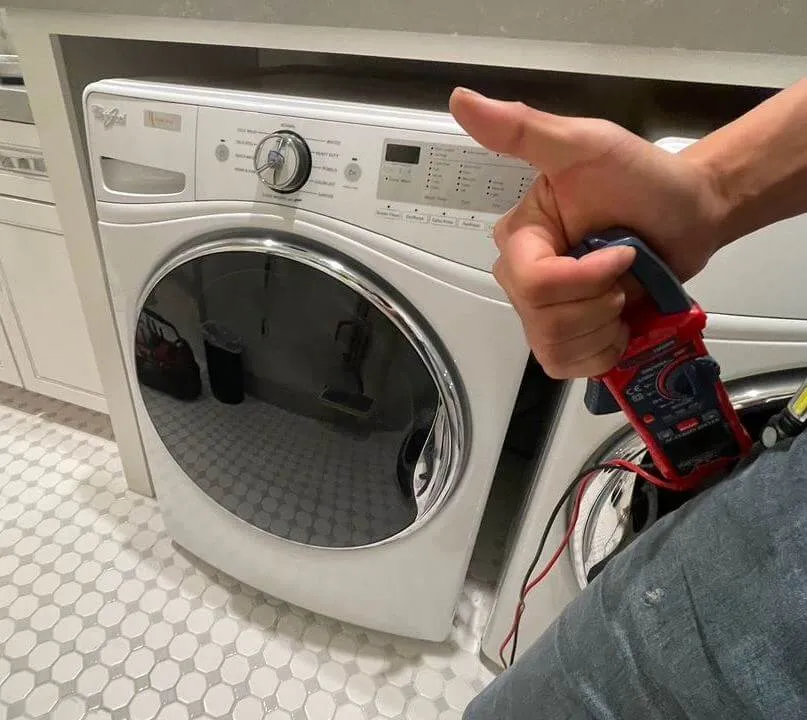 Front-loading white washing machine in a laundry room with a person holding a multimeter giving a thumbs up.