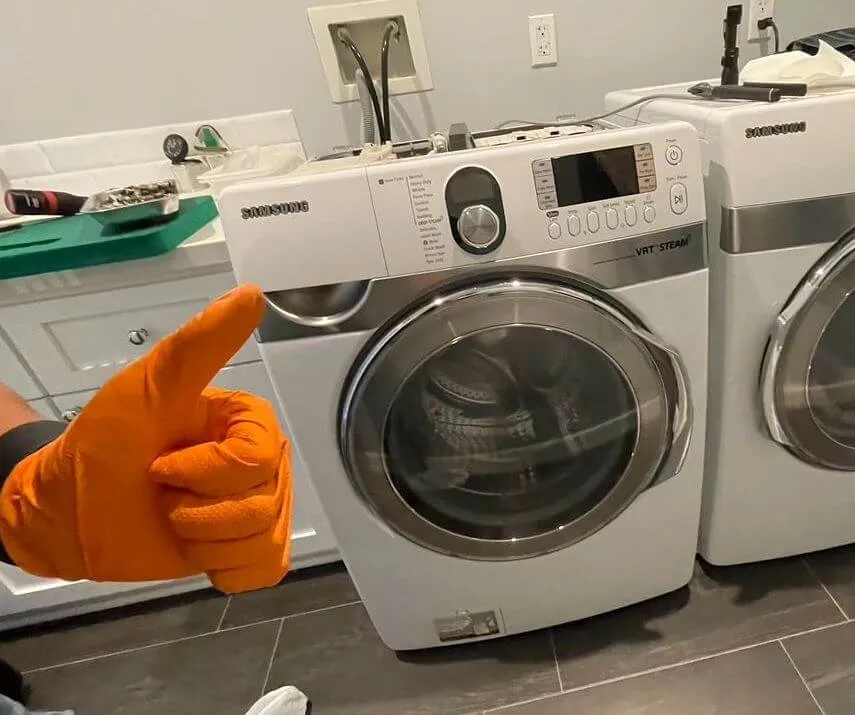 Front-loading Samsung washing machine and dryer in a laundry room with a person giving a thumbs-up wearing an orange glove.