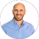 Smiling bald man with a beard wearing a light blue button-up shirt against a white background.