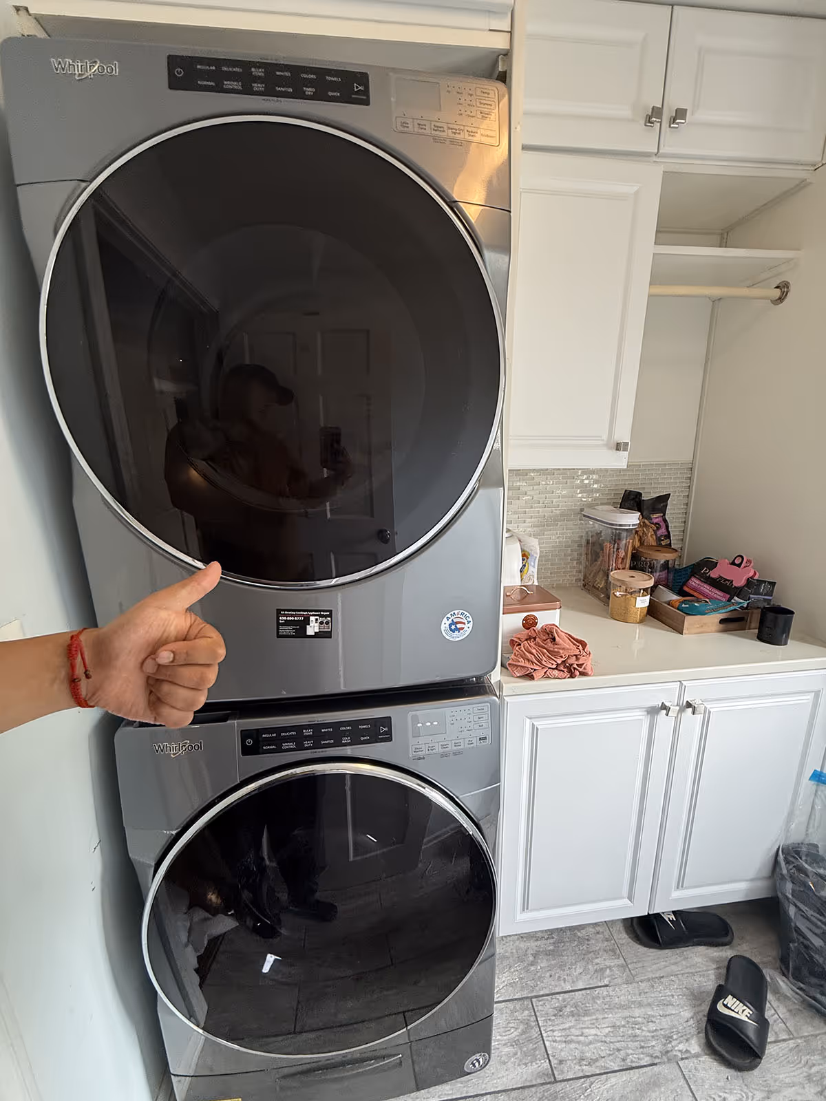 Stacked Whirlpool washer and dryer units in a laundry room with white cabinets and tiled floor, a hand giving thumbs-up in front.