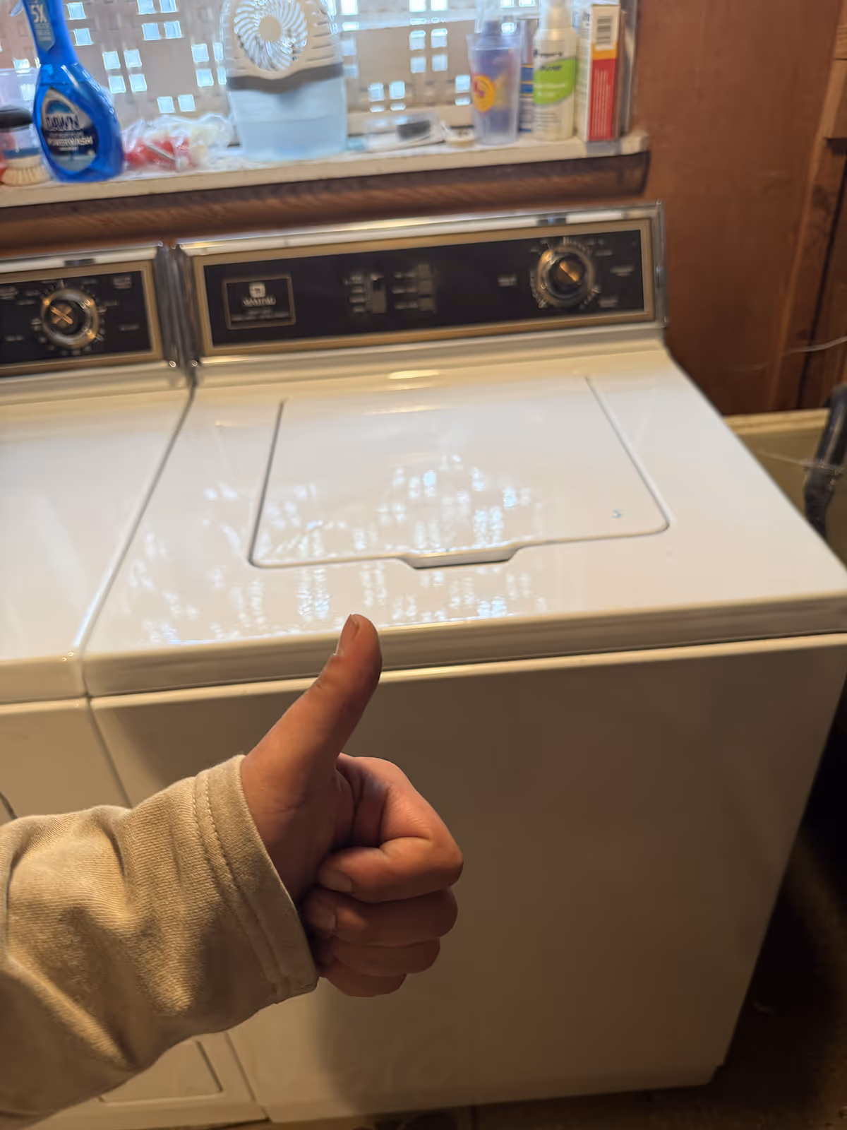 Hand showing thumbs up in front of a white top-loading washing machine in a laundry room.